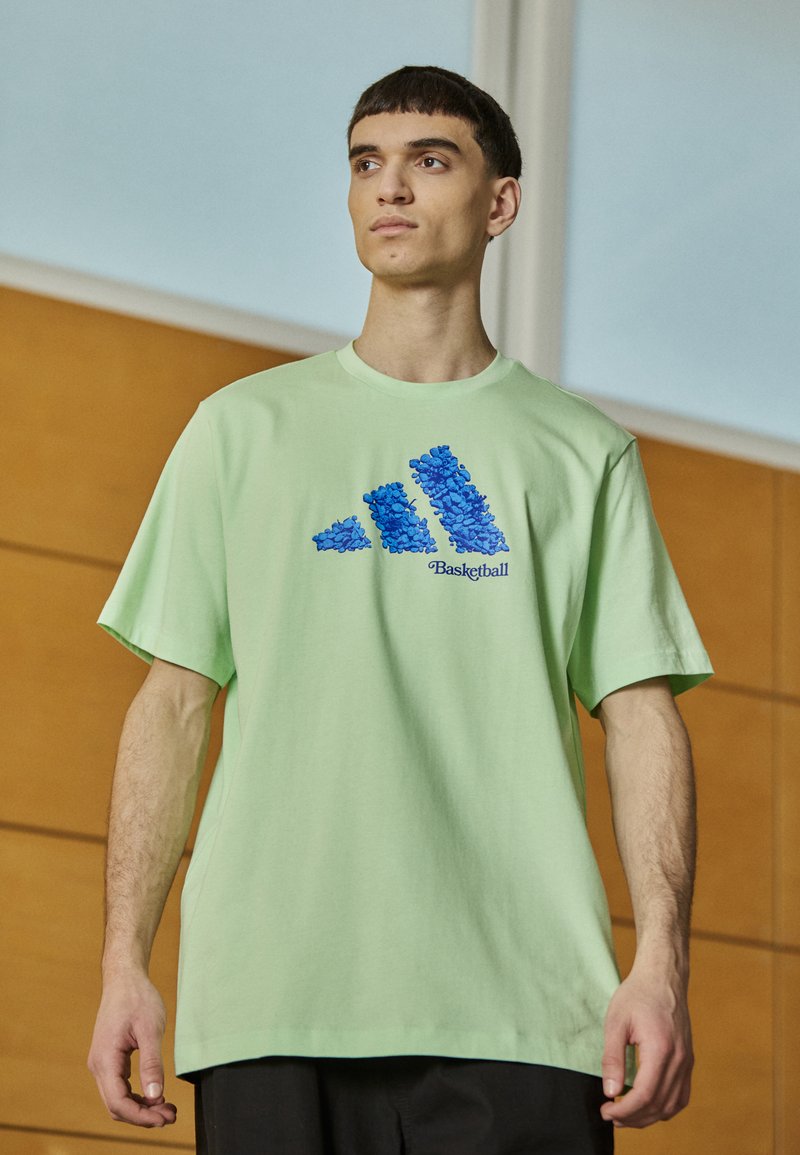 T-shirt oversize verde chiaro in cotone, con un design grafico blu di tre strisce e la parola "Basketball" sottostante.