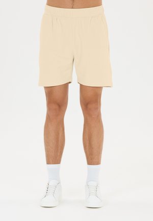 Mand iført beige shorts, hvide sokker og hvide sneakers, stående mod en ensfarvet hvid baggrund.