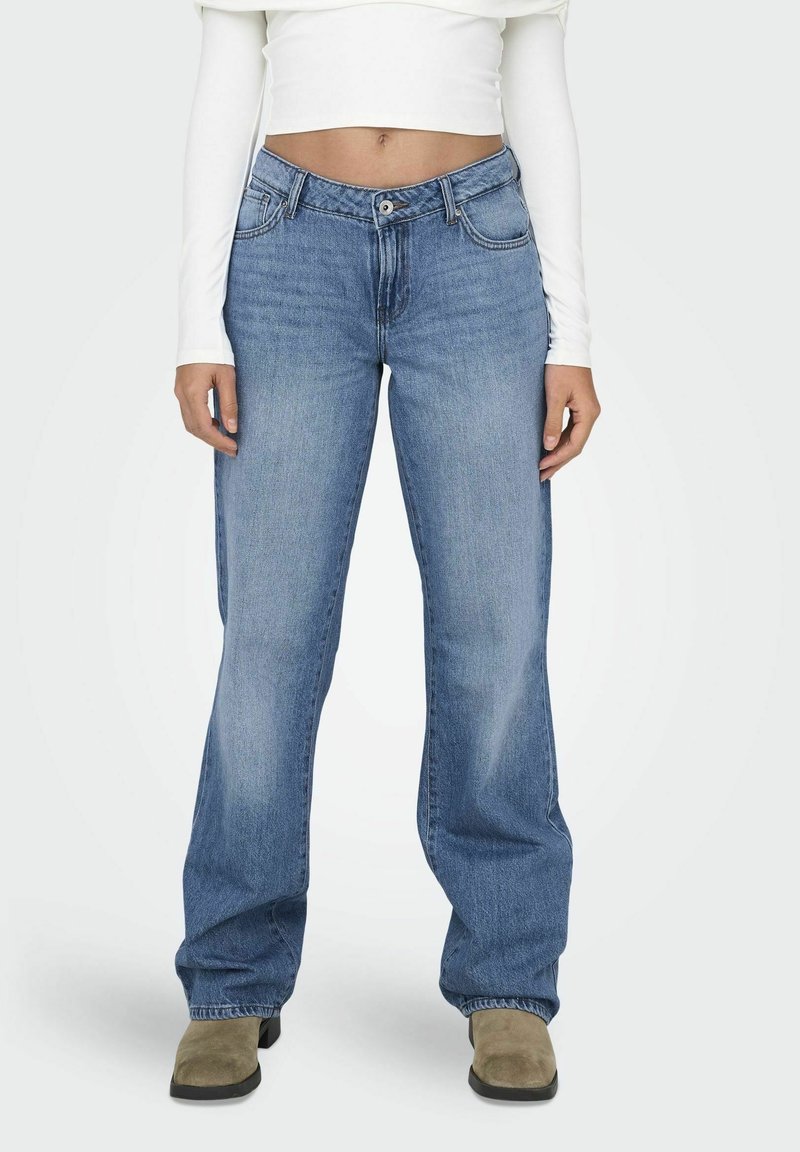 Femme portant un jean bleu clair à jambes larges, un crop top blanc à manches longues et des bottines beiges, debout devant un fond uni.