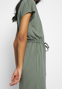 Mujer vistiendo un vestido corto de manga corta en color verde olivo con cintura ajustable, de pie de lado con el brazo izquierdo relajado a su lado.