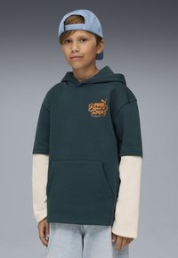 Felpa verde scuro con maniche corte, logo arancione e tasca frontale, indossata sopra maniche lunghe bianche. Cappellino azzurro chiaro. Jeans di denim chiaro.