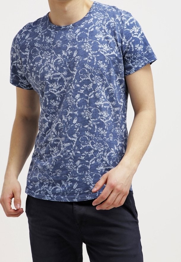 Homme portant un t-shirt bleu avec un motif floral blanc et un pantalon foncé, debout devant un fond clair uni.