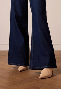 Jeans a gamba larga in denim scuro con dettagli di cucitura visibili, abbinati a tacchi a punta beige chiaro su un pavimento in legno.