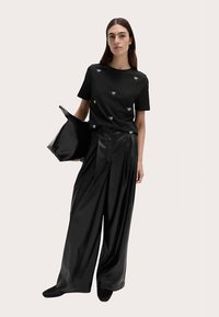 Czarna koszula z krótkim rękawem ozdobiona małymi białymi kokardkami, zestawiona z czarnymi szerokimi spodniami z faux skóry o wysokim stanie oraz elegancką czarną torbą typu tote.