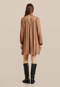 Vestido marrón a la altura de la rodilla con mangas largas, tejido texturizado, detalle de pliegues en la parte posterior y abertura en forma de ojo de cerradura. Lleva botas negras hasta la rodilla.