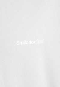 T-shirt bianco in cotone con il logo "Smilodox Sport" in testo stampato morbido. Il tessuto è liscio con una leggera texture.
