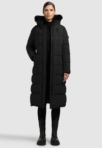 Zwarte lange pufferjas met een bont gevoerde capuchon, gequilt design en ritszakken. Gedragen over een zwarte trui en gecombineerd met zwarte kniehoge laarzen.