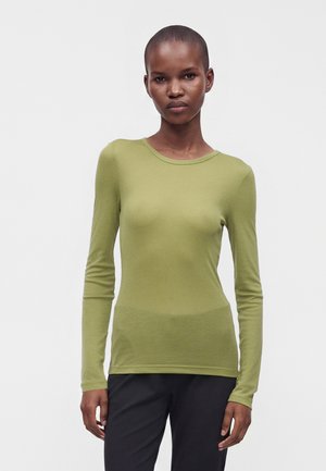 Femme portant une chemise à manches longues vert olive et un pantalon noir, debout devant un fond gris clair uni, regardant devant elle.