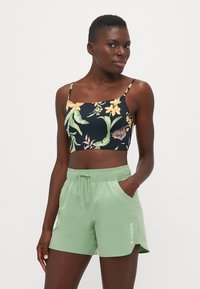 Svart crop top med blomstermønster, grønne lette shorts med elastisk liv og logo. Modell iført shorts med hendene i lommene.