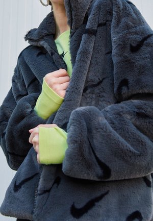 Personne portant une veste en fausse fourrure gris foncé avec des logos Nike noirs, partiellement zippée sur un sweat Nike vert clair.