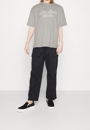 Camiseta gris de algodón con el gráfico "Low Lights Studios", combinada con pantalones cargo negros con bolsillos laterales y zapatos negros de ante sin cordones.