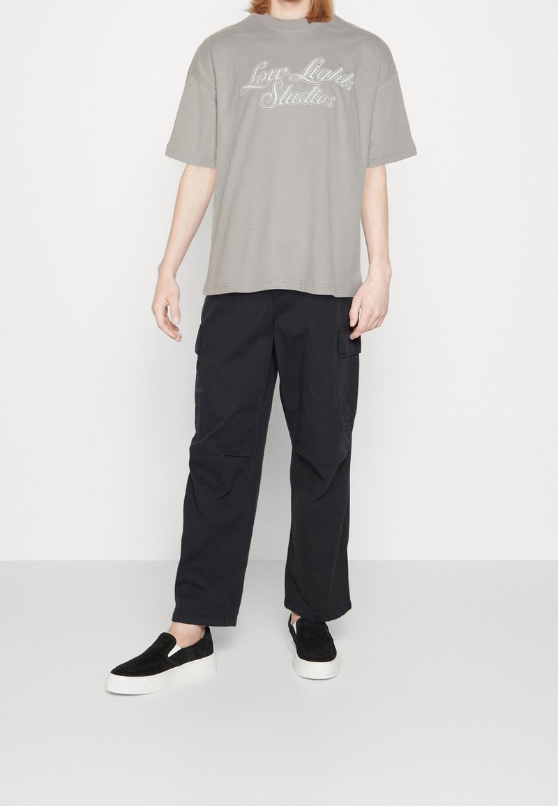T-shirt en coton gris avec le logo "Low Lights Studios", associé à un pantalon cargo noir avec poches latérales, et des chaussures slip-on en daim noir.