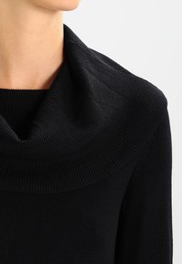 Sort strikket sweater med en bred, draperet cowl-udskæring og ribbet tekstur; nærbillede der fremhæver stoffets blødhed og struktur.