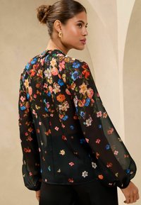 Blusa floreale in tessuto trasparente nero, con fiori colorati e farfalle, maniche lunghe e gonfie, e scollatura arricciata.
