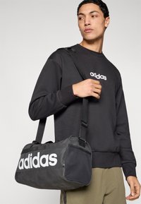 Melns duffle tipa soma ar baltu "adidas" logotipu, izgatavota no izturīga materiāla; tai ir teksturēts paneļa un regulējams plecu siksna.