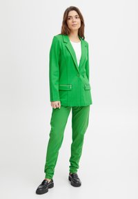 Blazer verde con cuello con muesca y dos bolsillos con cremallera, acompañado de pantalones verdes a juego. Tejido suave y corte ajustado. Mocasines negros.