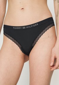 Crne tange s čipkastim rubom, s širokim pojasom na kojem je oznaka "TOMMY HILFIGER" u metalik koncu. Mekana tekstura tkanine.