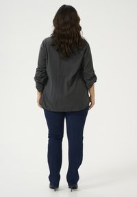 Chemise à manches longues gris foncé avec les manches retroussées, associée à un jean slim-fit bleu foncé et des chaussures à talons noires, vue de derrière.