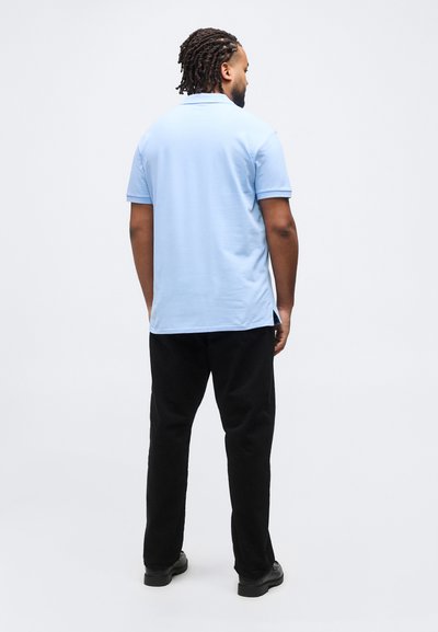 Άνδρας με πλεγμένα μαλλιά που φοράει ανοιχτό μπλε κοντομάνικο πόλο πουκάμισο, μαύρο παντελόνι και μαύρα παπούτσια, στέκεται γυρισμένος με την πλάτη σε λευκό φόντο.