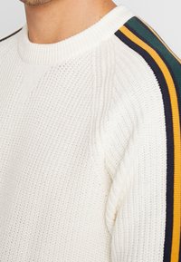 Sweter dziany w kremowym kolorze z prążkowaną fakturą, z paskami w kolorach zielonym, granatowym i żółtym wzdłuż szwów na ramionach.