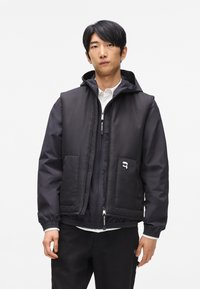 Melna kapucēta jaka ar priekšējiem kabatām, quilted tekstūra un r�āvējslēdzējs. Iespējas logotipa detaļa uz kabatas un elastīgas aproces.