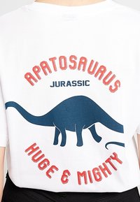 Valkoinen t-paita, jossa on sininen Apatosaurus-graafikka ja punaiset tekstit: "APATOSAURUS," "JURASSIC," ja "HUGE & MIGHTY" lihavassa typografiassa.
