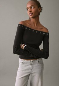 Crna bluzica bez ramena s srebrnim rupicama, dugim rukavima i prilagođenim dizajnom, u kombinaciji s svijetlosivim trapericama visokog struka.