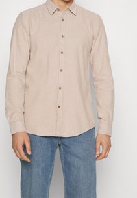 Jasnoróżowa koszula na guziki z teksturowanej tkaniny, z kołnierzem typu „klapka”, brązowymi guzikami, długimi rękawami i zaokrąglonym dołem.