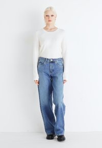 Lightweight, long-sleeve white sweater paired with high-waisted, loose-fit blue denim jeans. Model wears black chunky-soled shoes.