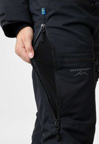 Zwarte waterdichte broek met ritsen en een mesh voering. Beschikt over een logo, een Zweedse vlag patch en meerdere ritszakken voor opslag.