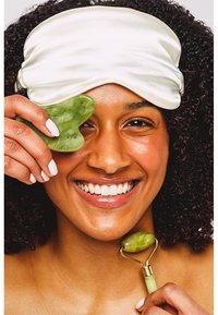Jade-Gesichtsroller und Gua Sha-Werkzeug aus grünem Stein; eine Seidenschlafmaske bedeckt die Stirn. Die Benutzerin hat lockiges Haar und zeigt ein lächelndes Gesicht.