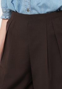 Brune skreddersydde bukser med folder, laget av glatt stoff, med en strukturert passform. Brukt med en lyseblå jeansskjorte.