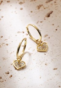 Orecchini a cerchio in oro con pendenti a forma di cuore, caratterizzati da scintillanti pietre trasparenti incastonate nel cuore. Finitura liscia e superficie lucida.