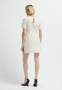 Vrouw met kort blond haar, gekleed in een crème jurk met korte mouwen, met gestreepte witte maillots en donkerbruine sleehakken, staand met haar rug naar de kijker gekeerd.