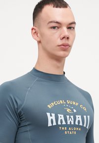 Dlhý rukáv rash guard v tmavo šedej farbe s grafickým dizajnom "Hawaii" v bielej a žltej farbe, vyrobený z pružného, hladkého materiálu.