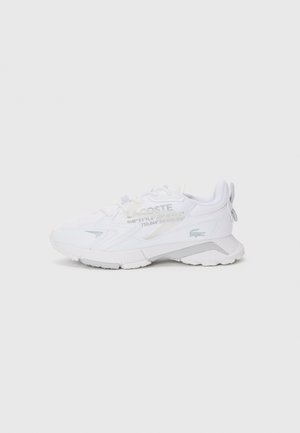Baskets blanches Lacoste avec semelle texturée, logo latéral et détails discrets de texte imprimé sur la tige, montrées de profil sur un fond uni.