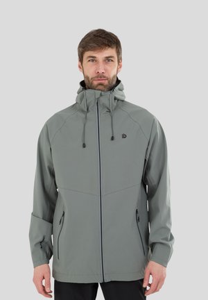 Uomo che indossa una giacca grigia con cappuccio e zip, con coulisse, in piedi davanti a uno sfondo semplice grigio chiaro.