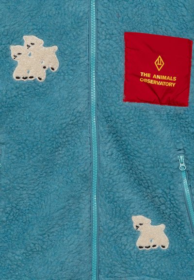 Giacca in pile azzurro chiaro con zip frontale, caratterizzata da due topolini polari beige e una tasca quadrata rossa etichettata "THE ANIMALS OBSERVATORY."