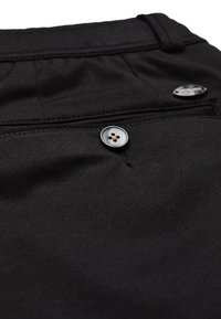 Schwarzer Stoff mit einer Taschenöffnung mit Knopf und einer Taillennaht, die ein kleines, markiertes Metalletikett in der oberen rechten Ecke zeigt.