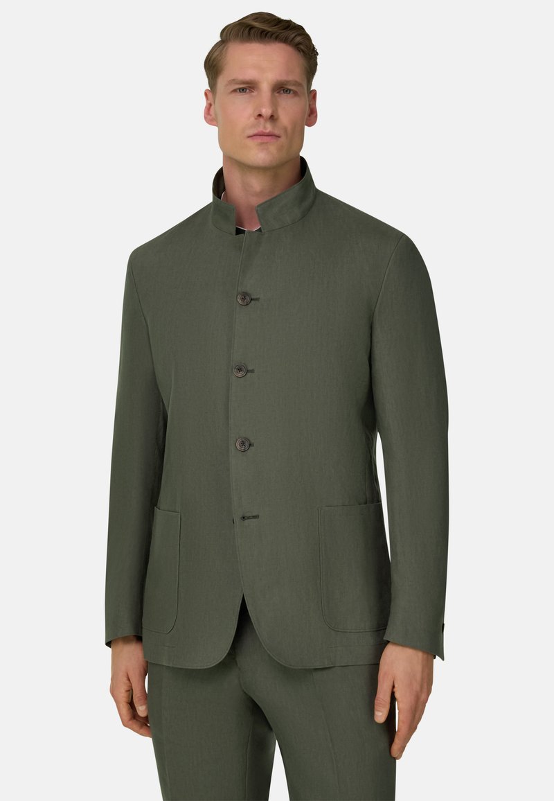 Homme portant un costume olive vert sur mesure avec un col montant et trois boutons, debout devant un fond clair uni.