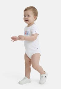 Παιδί σε όρθια θέση που περπατάει, φορώντας λευκό φορμάκι με το λογότυπο "Tommy" και λευκά αθλητικά παπούτσια, μπροστά σε απλό λευκό φόντο.