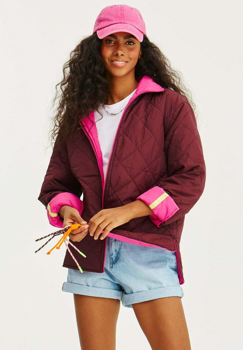 Prošívaná burgundská bunda s růžovými akcenty, zipy a manžetami. V kombinaci s džínovými šortkami a světle růžovou čepicí. Drží barevné brčka.