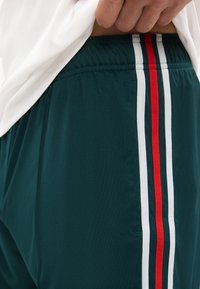 Κοντινό πλάνο ενός ατόμου που σηκώνει ένα λευκό πουκάμισο, αποκαλύπτοντας σκούρα πράσινα αθλητικά σορτς με κόκκινες και λευκές κάθετες ρίγες στο πλάι.