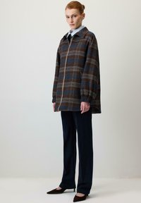 Rjava in modra karo jakna z visokim ovratnikom, zadrgo spredaj in nabranimi rokavi, kombinirana z mornarsko modrimi hlačami in temnimi koničastimi čevlji.