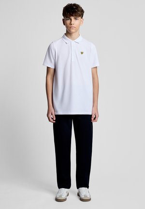 Teenager steht frontal und trägt ein weißes Poloshirt mit gelbem Logo, schwarze Hose und weiße Sneakers vor einem einfarbigen grauen Hintergrund.