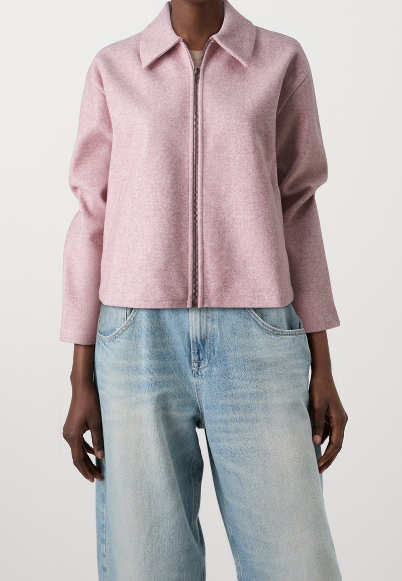 Personne portant une veste zippée rose clair avec un col et un jean taille haute en denim bleu clair, debout devant un fond uni.