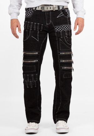 Mann trägt schwarze Hose mit mehreren Reißverschlüssen und weißen Nähten, weißen Gürtel, weiße Schuhe und grauen Pullover, sichtbar an der Taille.