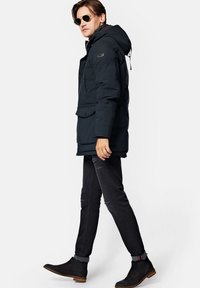 Ciemnoniebieska kurtka puchowa z kapturem, z przednią kieszenią i zapięciem na zamek. Noszona z wąskimi czarnymi dżinsami i botkami za kostkę.