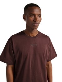 Jonge man met kort haar en oorbel, die een effen donkerbruine T-shirt draagt met de tekst "HALO" op de borst, kijkt iets naar rechts op een witte achtergrond.