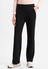 Schwarze, gerade geschnittene Hosen mit weicher Textur, einem breiten Bund und seitlichen Taschen, kombiniert mit hellen Sportschuhen.